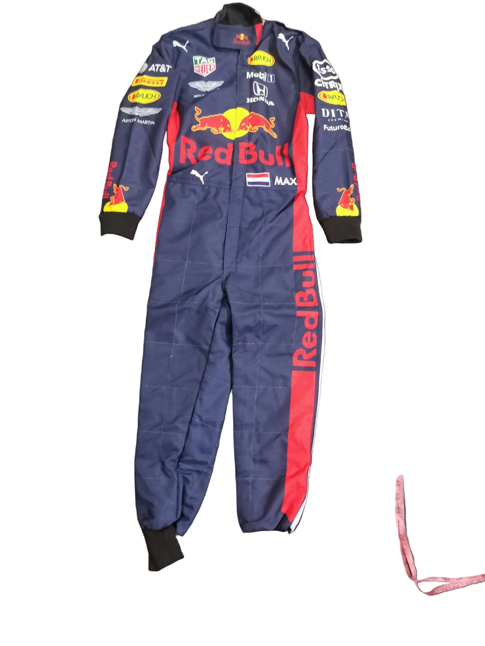 A navy blue Red Bull racing one-piece suit with red and white side stripes and multiple sponsor logos, laid flat on a dark gray fabric background next to a pink measuring tape.