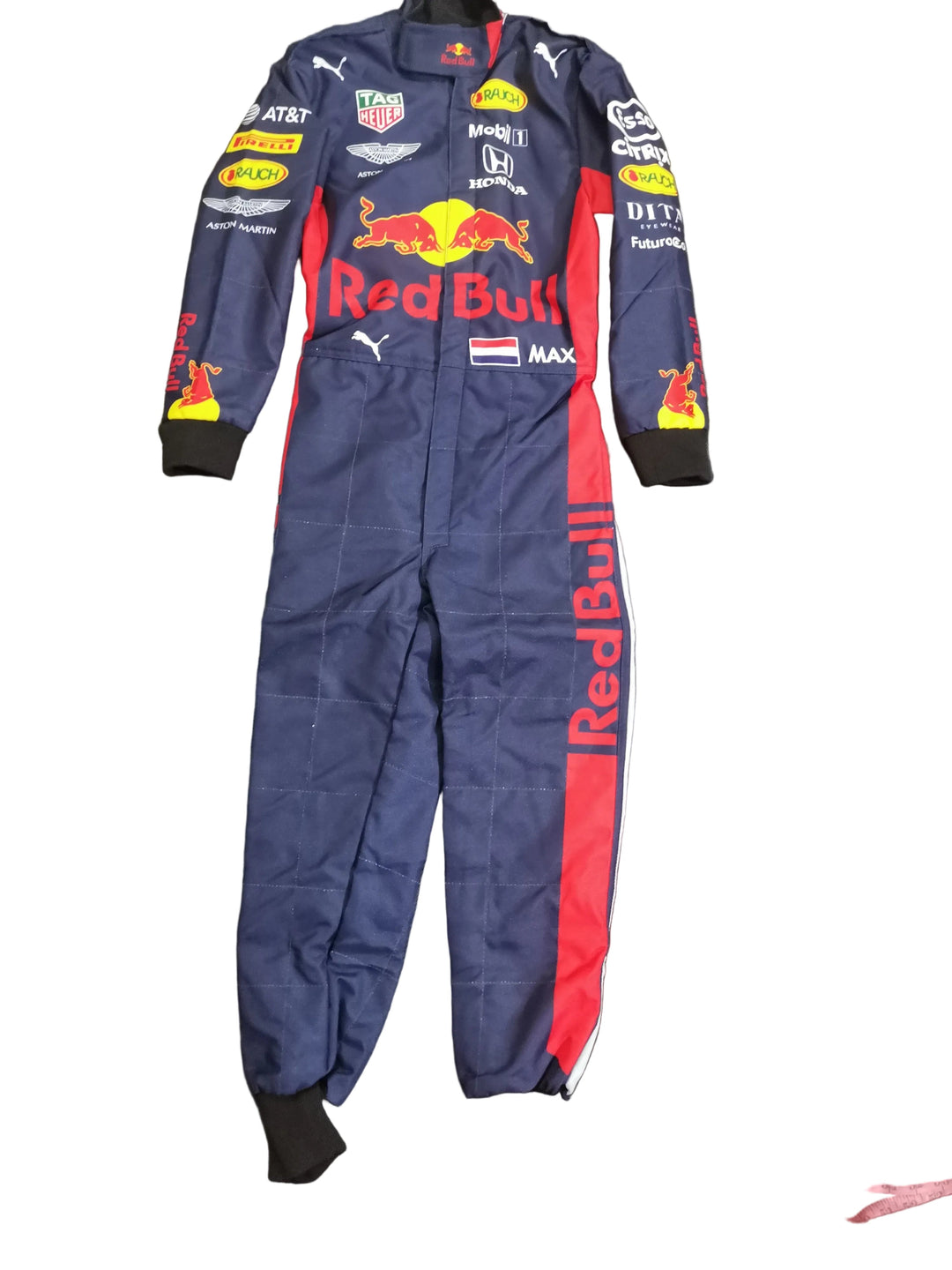 A full-length navy Red Bull racing suit laid flat on a dark gray fabric surface, with a pink measuring tape running vertically along the right side. The suit features multiple sponsor logos including TAG Heuer, AT&T, and Maersk, and a large Red Bull logo on the chest.