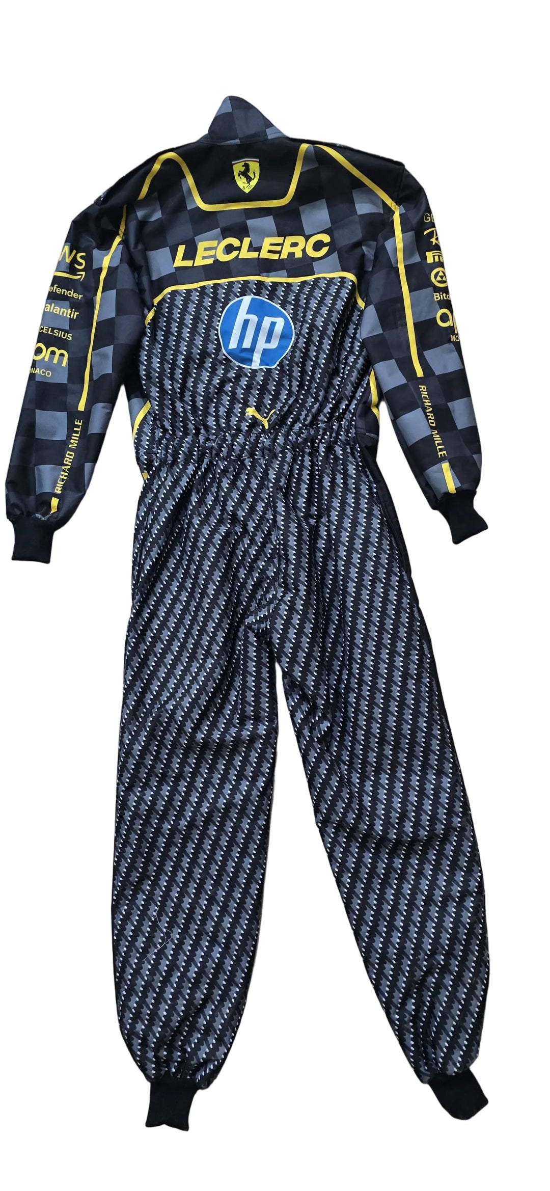 A full-body racing suit laid flat on a beige tiled floor. The suit is black with a gray and white geometric pattern on the lower half, a black and gray checkered pattern on the upper half and sleeves, and yellow accents.