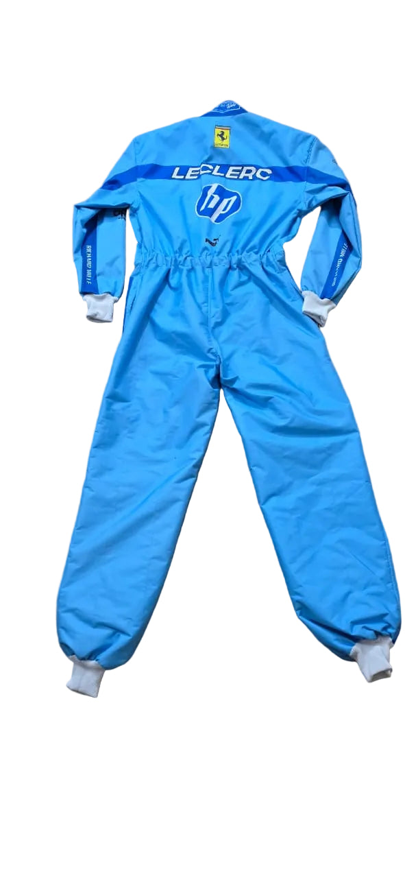 A light blue LeManc HP racing one-piece suit with white cuffs, laid flat on a beige tiled floor. The suit features 'LE MANC' and the HP logo on the back, along with a small yellow Ferrari logo near the neck.