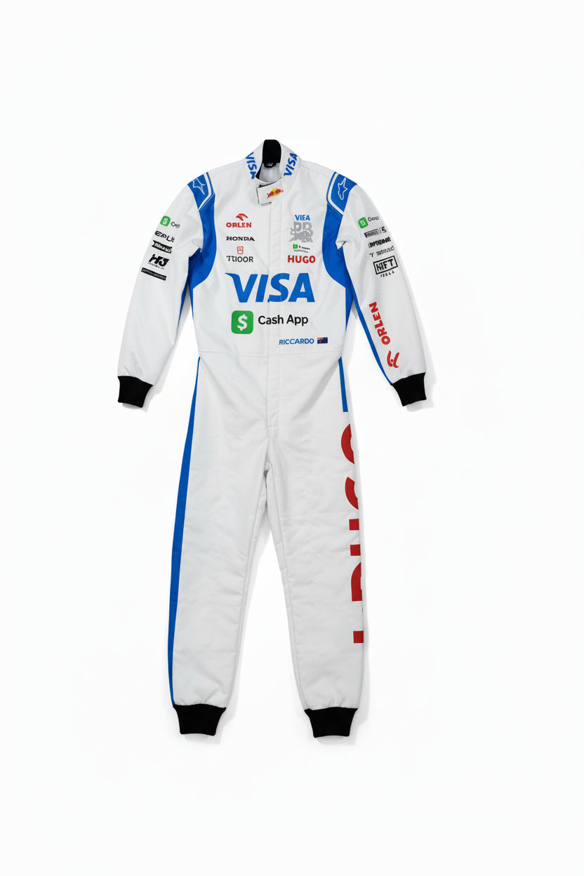 A white racing one-piece suit with blue and red sponsor logos, laid flat on a brown concrete floor. A bare foot is visible at the bottom left corner.