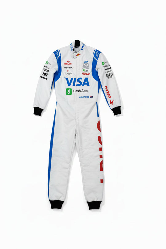 A white racing one-piece suit with blue and red sponsor logos, laid flat on a brown concrete floor. A bare foot is visible at the bottom left corner.