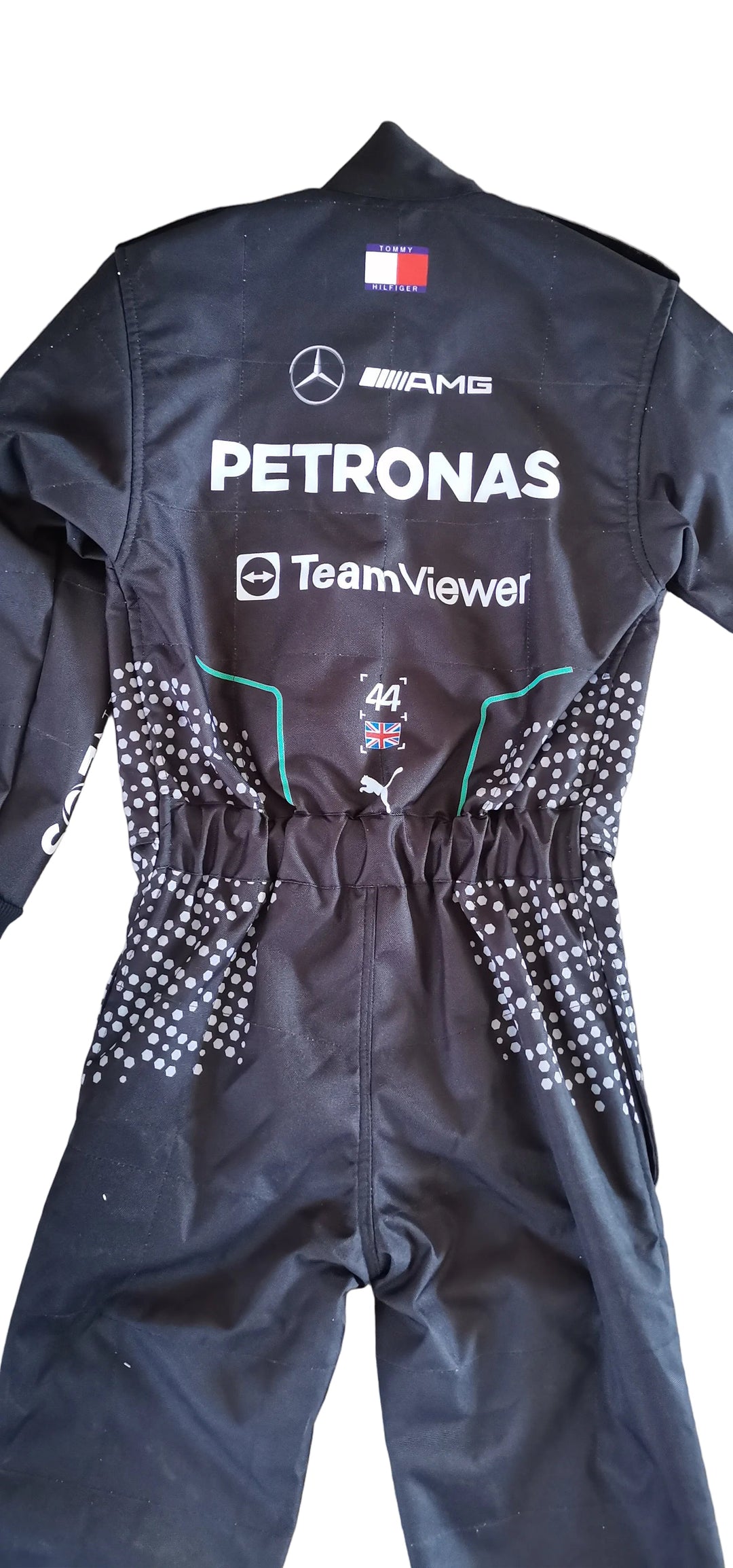A black Mercedes-AMG Petronas Formula 1 racing one-piece jumpsuit, shown from the back, laid flat on a light-colored wooden floor. The suit features white sponsor logos, a small French flag patch, and white rhinestone embellishments on the sides.