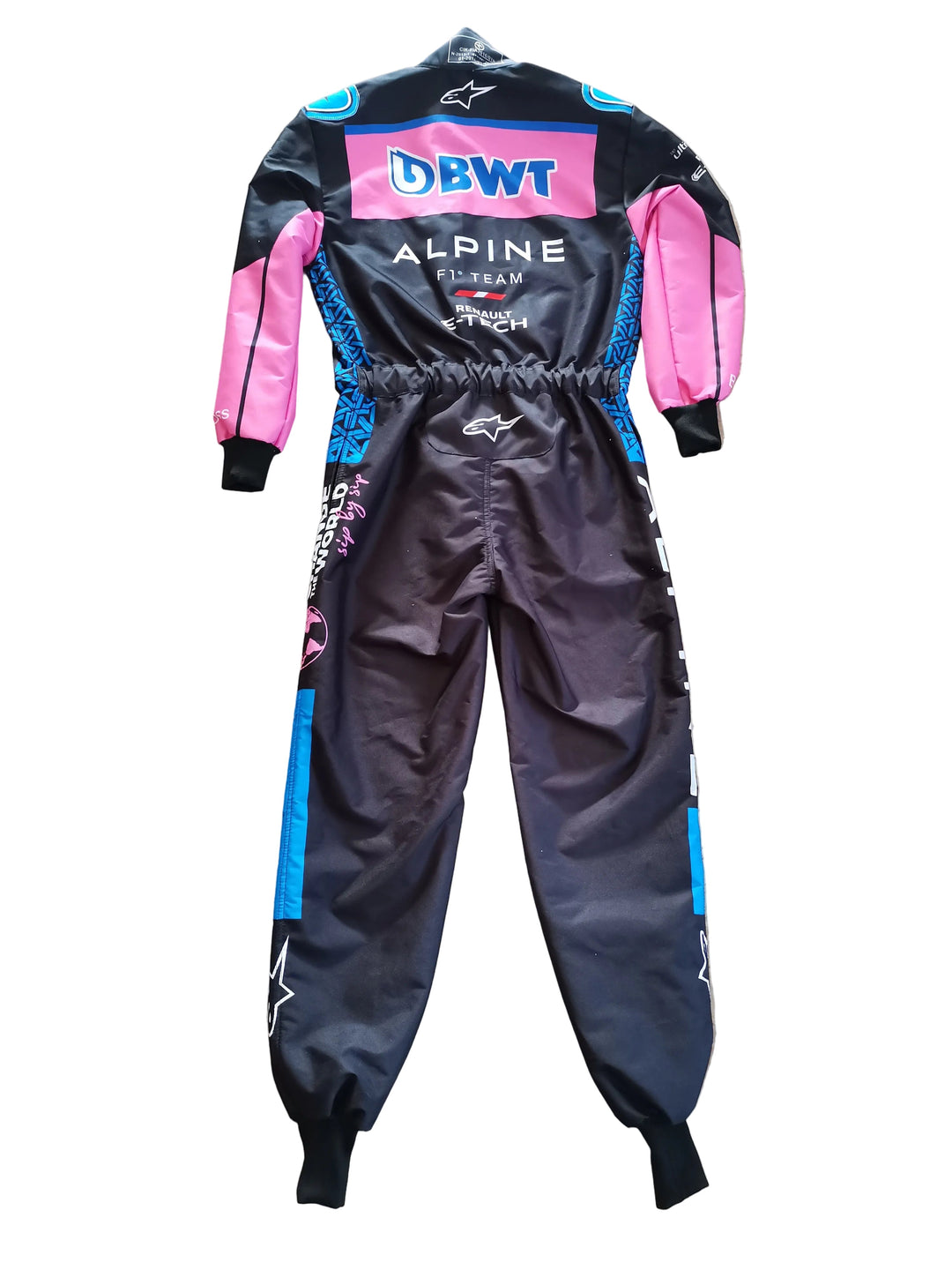 A full-body Alpine F1 team racing suit by Alpinestars, laid flat on a beige marble floor. The suit features a black base with vibrant pink sleeves and side panels, blue accents, and white branding.