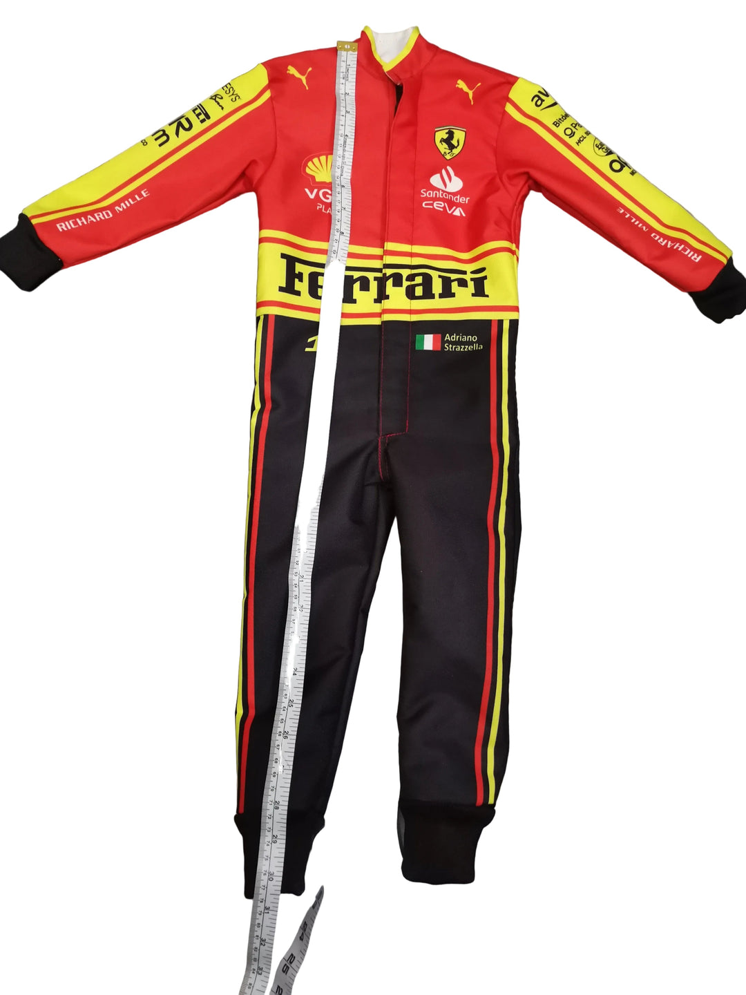 A Ferrari one-piece racing suit laid flat on a gray tiled floor, with a measuring tape running vertically down the center to show the height. The suit is red and yellow on the upper body and sleeves, and black on the lower body, with Ferrari, Puma, and Shell logos visible.