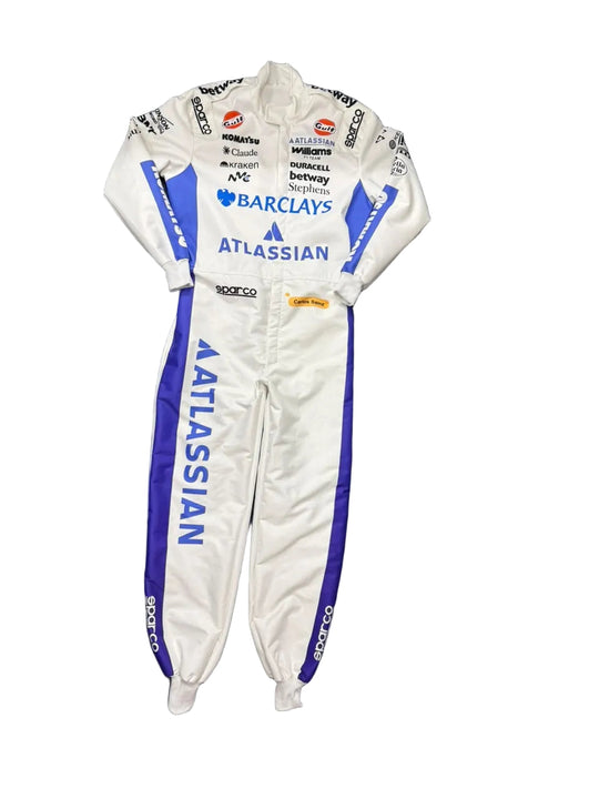 A white and blue ATLASSIAN racing suit laid flat on a dark blue blanket, with visible sponsor logos including Barclays and Soco. The photo is taken in a casual indoor setting with floor tiles, shoes, and storage items visible around it.