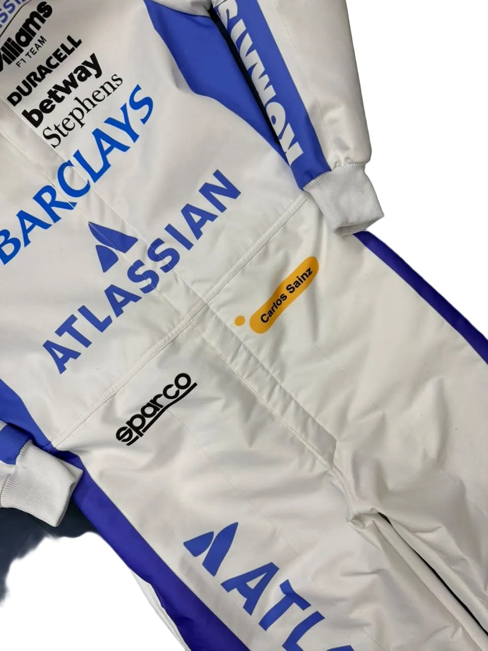 A close-up of a white racing suit with blue and purple panels, featuring sponsor logos including Barclays, Atlassian, Sparco, Williams, and Duracell.