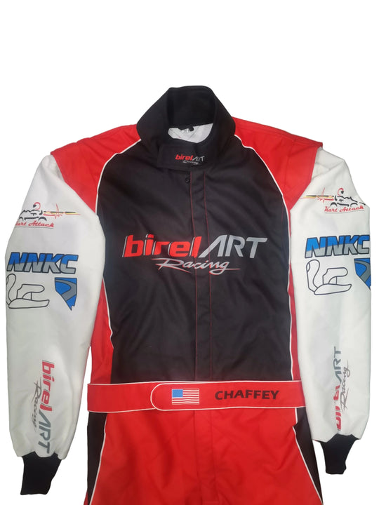 A full-body racing suit laid flat on a gray tiled floor. The suit is primarily black on the torso, with red side panels and white sleeves.