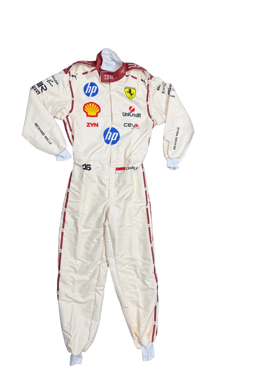 A cream-colored Scuderia Ferrari F1 racing suit with red and white trim and various sponsor logos, laid flat on a dark blue pillow over a gray tiled floor.