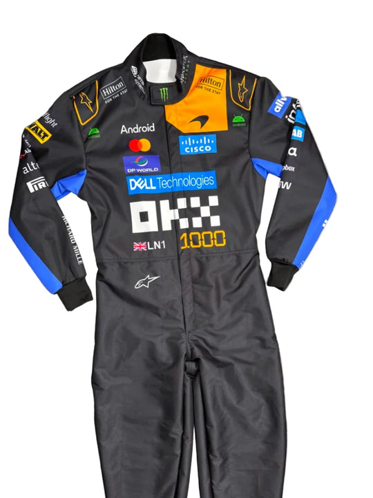 A black and blue Alpinestars racing suit with various sponsor logos, laid flat on a gray tiled floor. A person's bare foot is visible in the top-left corner.