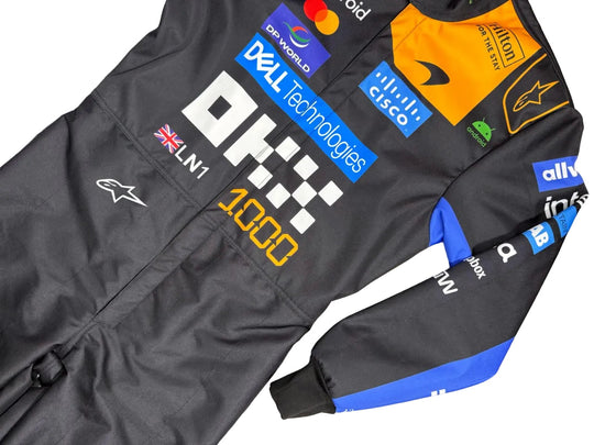 A black Alpinestars F1 racing suit with various sponsor logos and a blue and orange stripe on the sleeve, laid flat on a gray tiled floor.