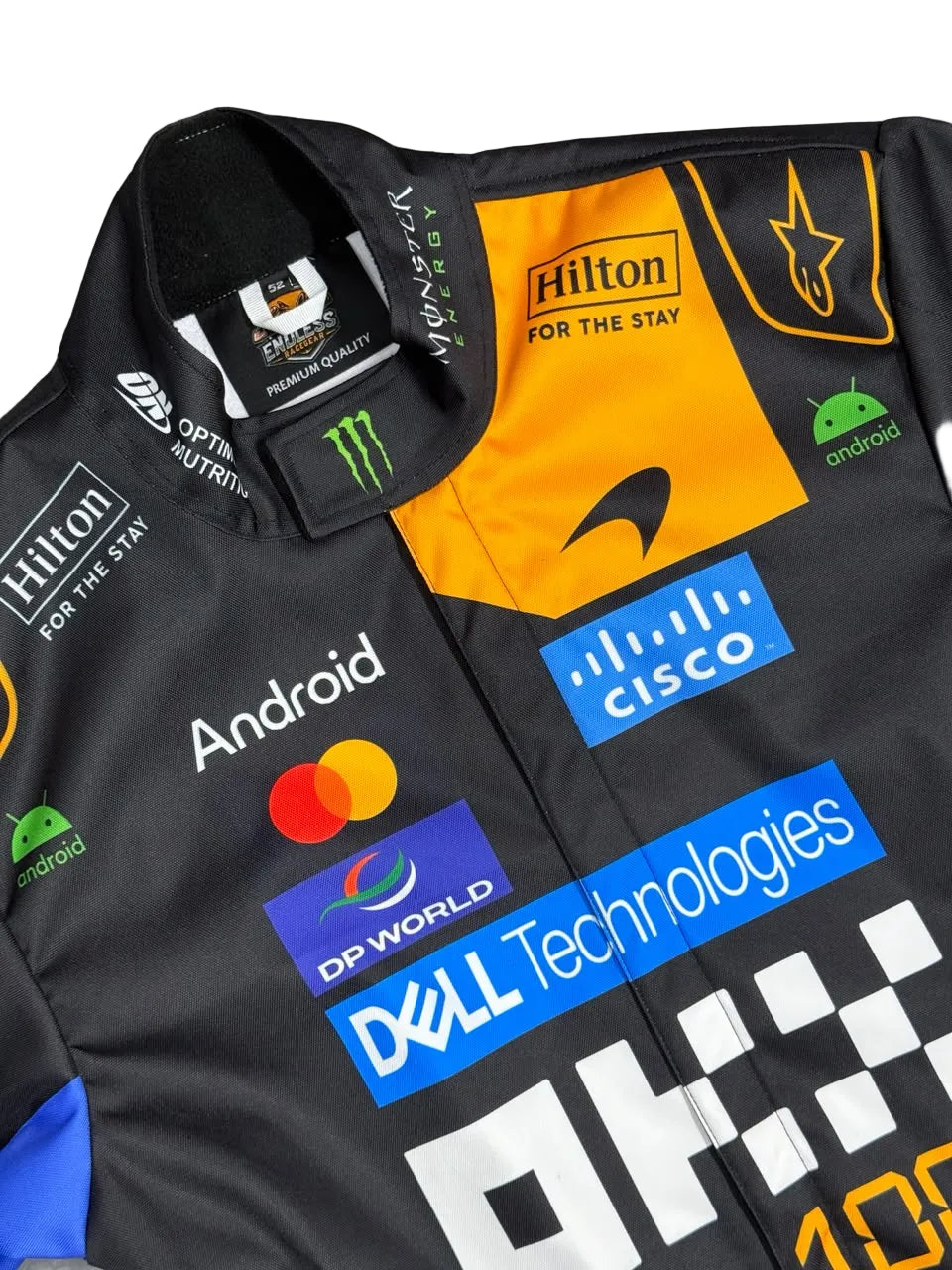 A close-up of a black racing team jacket featuring prominent sponsor logos including Hilton, Android, Mastercard, Cisco, Dell Technologies, and DP World, with a bright orange chest panel and a black collar.