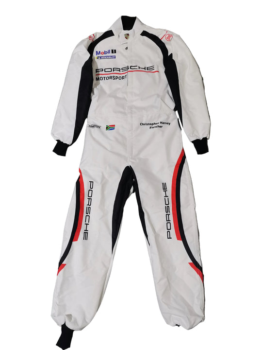 A white Porsche Motorsport racing suit with black and red side panels, laid flat on a gray marble-patterned tile floor. The suit features various sponsor logos including Mobil 1, Mobil 1 Racing, and Christian de Sousa Racing.