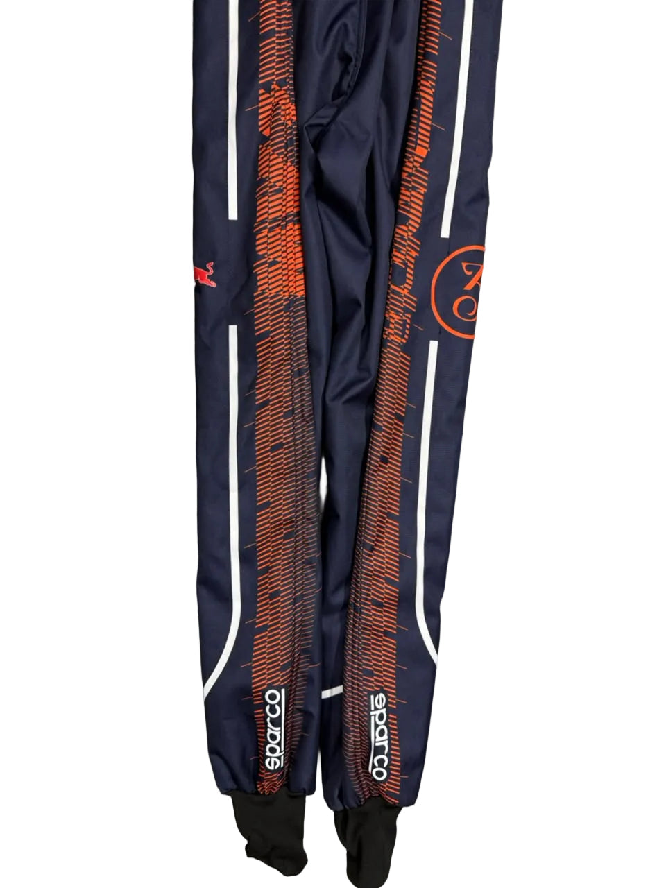 A pair of navy blue Sparco track suits laid flat on a gray marble tile floor. The suits feature orange and black patterned side stripes, white side piping, and black leg warmers attached at the top.